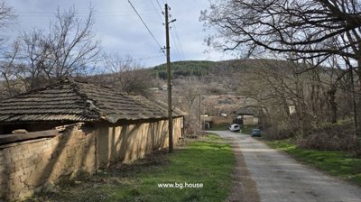 Narrow village road bordered by a stone wall and leafless trees, with a hill and scattered houses in the background