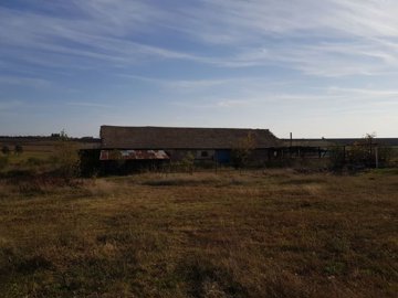 Wide view of the farm building with tiled roof and metal fencing, set in open grassy land under a partly cloudy sky.