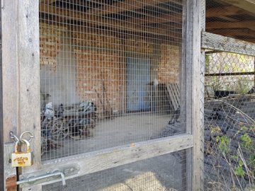 Close-up of a wooden enclosure with wire mesh and a rusty padlock, with the farm building visible behind.
