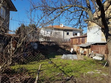 Fenced grassy land with garden beds covered in plastic and bare trees near residential buildings in Popovo