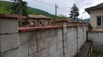 Concrete block boundary wall topped with red tiles, enclosing a yard with traditional village houses and forested hills in the distance.