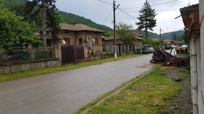 Street view showing village houses with stone boundary walls, metal fences, and a woodpile stacked beside the road.