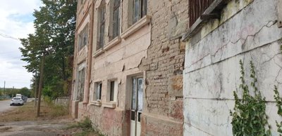 View of an old stone building showing peeling plaster and cracked walls along a roadside with parked cars and trees.