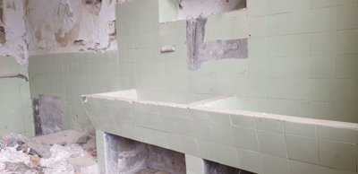 Room showing green wall tiles, damaged plaster, and scattered debris on the floor