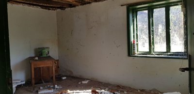 An empty room featuring exposed wooden ceiling beams, white walls, a green-framed window, and scattered debris on the floor.