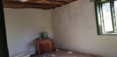 An empty room showing exposed wooden ceiling beams, white walls, a green-framed window, and debris scattered on the floor.
