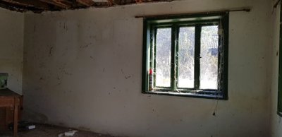 Empty room with exposed wooden ceiling beams, white walls, a green-framed window, and debris on the floor.