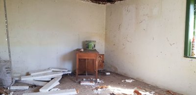 An empty room featuring exposed wooden ceiling beams, white walls, a green-framed window, and scattered debris on the floor.