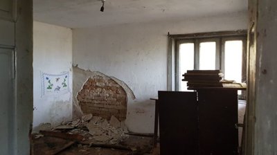 Interior of a room featuring white walls with damaged plaster and stacked wooden boards near the window.