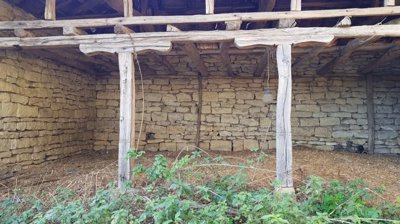 Exterior view of a stone outbuilding featuring wooden beams and an open side, with green plants growing nearby.