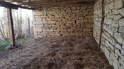 View inside a partially enclosed stone building showing wooden beams and straw covering the floor.