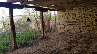 Stone outbuilding interior with wooden beams overhead and straw covering the ground, with vegetation visible outside.