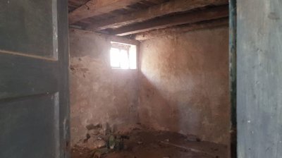 Interior view of a small room showing rough plaster walls, wooden ceiling beams, and a small window letting in light.