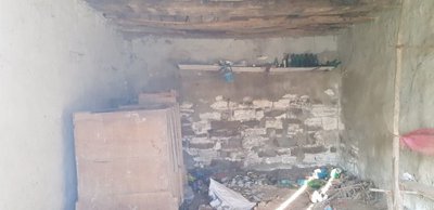 Rustic interior room showing stone and plaster walls, wooden ceiling beams, and various scattered items on the floor and shelves.