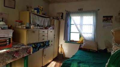 Room interior with a cupboard, bed covered with green blanket, and window with lace curtains.