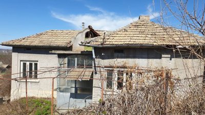 Side perspective of a grey village house featuring a tiled roof and an enclosed porch, with dry vegetation and bare branches around the property.