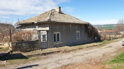 Rear side of a traditional grey village house with a tiled roof and several windows, located next to a dirt road and open landscape.