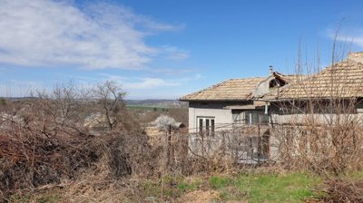 Village house situated on a corner plot, featuring a stone wall and a dirt road running alongside the property.