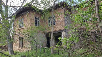 Two-storey rural house made of brick and stone with multiple windows, partially obscured by trees