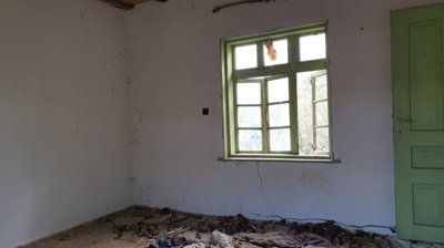 Empty interior room with green window and door frames, white walls, wooden ceiling, and cracked floor