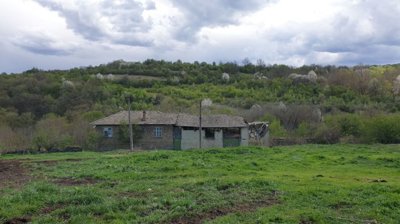 View of a traditional rural house with a tiled roof set against a backdrop of green hills and trees under a cloudy sky.