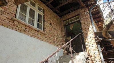 Brick wall featuring two white-framed windows and a metal door at the top of concrete stairs with rusty railing.