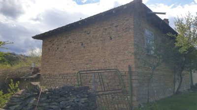 Corner view of a stone and brick house with small trees and grass around, and a stone fence in the foreground.