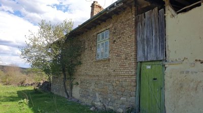 Stone and brick wall of the house with windows and a green door, surrounded by grass and trees.