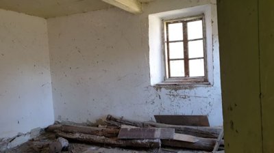 Interior view of an old room showing white plaster walls, wooden ceiling beams, and a small window with wooden frame.