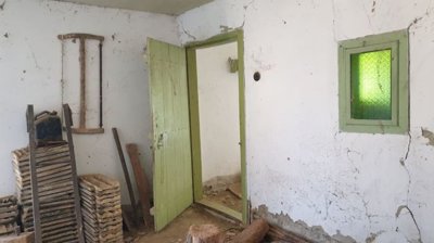 Interior room featuring a green wooden door, a small green window, stacks of wooden tiles, and farming tools leaning against the wall.