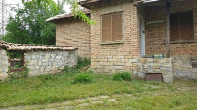 Exterior view of a small brick house with wooden shutters and stone base, set in a grassy yard with trees nearby.