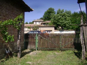 View of a wooden gate and fence beside a brick house wall, with trees and clear blue sky in the background.