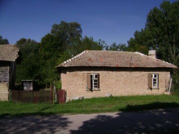 Side view of a single-storey brick house featuring a tiled roof and wooden window shutters, with green grass and trees around.