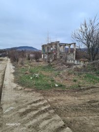 A ruined two-story concrete building frame stands on a grassy plot with bare trees and a dirt road beside it under a cloudy sky.