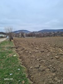 Ploughed farmland adjacent to a dirt road where a car is parked, with village houses and hills visible in the background.