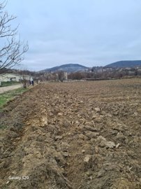 Ploughed field with leafless trees and village houses on hills visible in the distance under a cloudy sky.