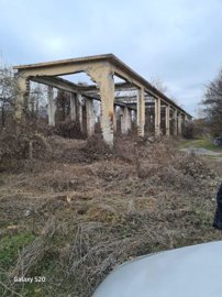 A long ruined concrete structure frame is surrounded by dry grass and bushes under a cloudy sky.