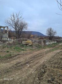 A dirt road with tire tracks runs alongside a low concrete wall with bare trees and hills in the background under a cloudy sky.
