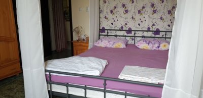 Bedroom showing a double bed with purple bedding, floral curtains, wooden wardrobe, and bedside table