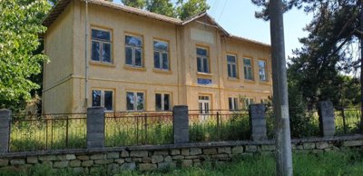 Exterior view of the two-storey former school building with yellow walls and many windows, set behind a stone and metal fence with overgrown grass
