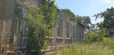 Exterior view of the former school building showing tall windows and an entrance partially covered by vegetation