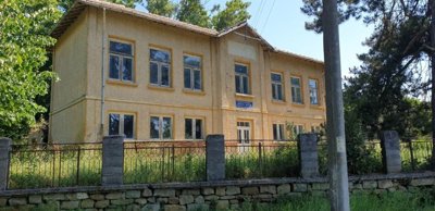 Front facade of the former school building showing symmetrical windows and entrance door behind a stone and metal fence