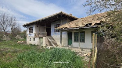 Side perspective of a Bulgarian house featuring stone basement, wooden balcony, tiled roof, and green doors and windows.