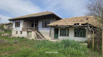 Front exterior of a traditional Bulgarian house featuring stone basement, wooden balcony, and garden with wild grass and flowers.