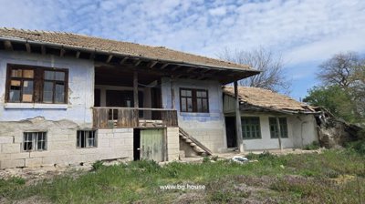 Side view of a traditional Bulgarian house showing stone basement, wooden balcony, green doors and windows, and tiled roof.