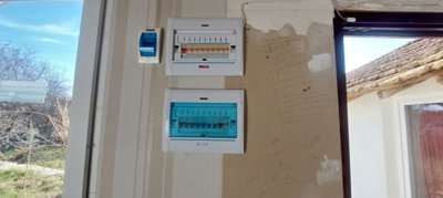 Two electrical fuse boxes installed on a wall with handwritten notes nearby and a window showing outdoor trees.