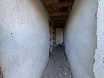 Interior view of a narrow corridor with unfinished plaster walls and exposed wooden ceiling beams.