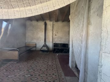 Interior room showing a single metal bed frame, a small sofa, a wood stove against a rough plaster wall, and patterned tile flooring.