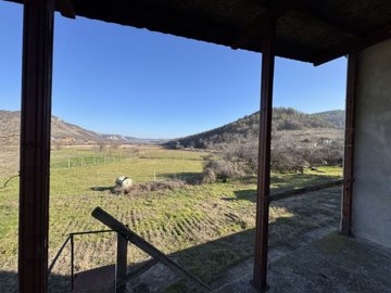 View from a covered balcony showing fenced grassy land, scattered trees, and hills in the distance under a clear blue sky.