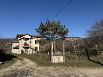 Rural house adjacent to a fenced yard with a pine tree, with hills visible in the background under clear sky.